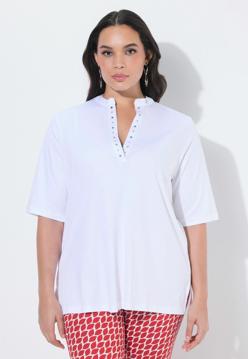 Kobieta o długich ciemnych włosach, ubrana w biały krótki top z dżetami na dekolcie w serek oraz czerwone spodnie z wzorem, stoi na jasnym tle.