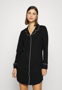 Vestido de noite preto, com fecho à frente e feito de tecido suave, com mangas longas e bainhas brancas nas golas e punhos. Barra arredondada.