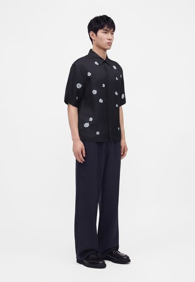 Homme debout portant une chemise noire à manches courtes avec des fleurs blanches, un pantalon large sombre et des chaussures noires sur fond blanc.