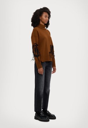 Brauner gestrickter Rollkragenpullover mit schwarzem gesticktem Detail und fransigen Akzenten, kombiniert mit dunklen grauen Jeans und klobigen schwarzen Schuhen.