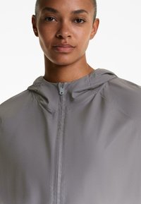 Giacca leggera grigia con cappuccio, dotata di una zip frontale e una superficie liscia. Vestibilità adatta senza modelli o accenti di design prominenti.