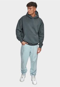 Grå hætte sweatshirt med en frontlomme, parret med lyseblå denimbukser og hvide sneakers. Glat tekstur og løs pasform.