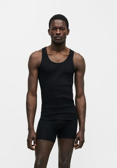 Άνδρας με σκουρόχρωμο δέρμα, που φοράει μαύρο αμάνικο τοπ και μαύρα στενά σορτς, στέκεται μπροστά σε ένα απλό ανοιχτό φόντο.