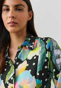 Vícebarevná halenka s velkými květinovými a puntíkovými vzory na černém pozadí. Měkká látka s límcem a knoflíkovými detaily.