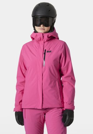 Rosa wasserdichte Skijacke mit Kapuze, taillierter Schnitt, Vorderreißverschluss und schwarzen Logodetails. Getragen mit schwarzen Handschuhen und einem Helm.