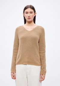 Beige strikkegenser med V-hals, med teksturerte mønstre og ribbestrikkede detaljer på mansjetter og nederkant. Brukt med lyse bukser.