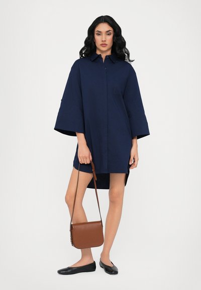 Abito camicia oversize blu navy con colletto, bottoni frontali e taschino sul petto; abbinato a ballerine nere e una borsa a tracolla marrone.