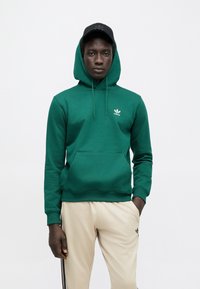 Zelená bavlnená mikina s klokankovým vreckom, nastaviteľnou kapucňou so šnúrkou a bielym logom Adidas na hrudi, v kombinácii s béžovými nohavicami.