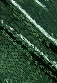 Grüner Paillettenstoff mit runden, glänzenden Pailletten, die in Reihen angeordnet sind. Die Textur ist glatt und reflektierend und erzeugt einen schimmernden Effekt.