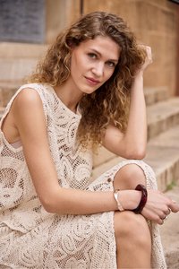 Femme aux cheveux bouclés en robe beige en crochet sans manches, assise sur des marches en pierre en extérieur, reposant son bras sur son genou, portant des bracelets argentés et foncés.