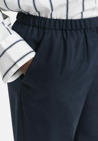 Hand in de zak van navyblauwe elastische taillebandbroek, gecombineerd met een witte longsleeve shirt met navy verticale strepen.