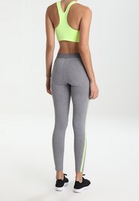 Mujer con un sujetador deportivo verde lima, leggins grises con rayas laterales verdes lima y zapatillas negras, de pie de espaldas sobre un fondo blanco.