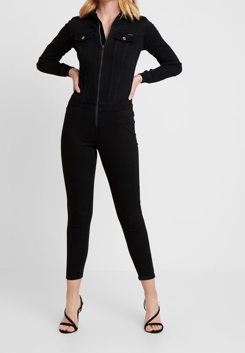 Black denim jumpsuit with long sleeves, zip front, two chest pockets, and slim-fit tapered legs. Paired with strappy black heels.