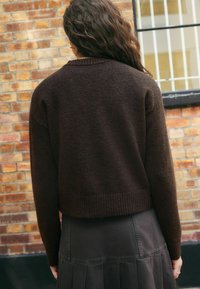 Brązowy sweter dzianinowy z ribbowanym dołem, noszony na ciemnej plisowanej spódnicy. Materiał ma miękką fakturę, charakteryzuje się luźnym krojem i opuszczonymi ramionami.