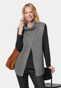 Grauer gestrickter ärmelloser Pullover mit einer Kragenform, der ein strukturiertes Muster und eine lockere Passform aufweist, kombiniert mit einem schwarzen Langarmoberteil und einer braunen Tasche.