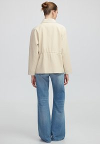 Beige textuur jasje met een ceintuur in de taille, kraag en lange mouwen, gecombineerd met lichtblauwe flare jeans.