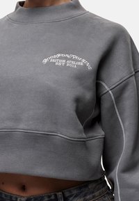 Felpa grigia corta con collo alto, caratterizzata da un logo bianco ricamato e una texture soffice. Include polsini e orlo a costine.