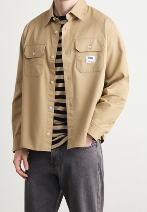 Chemise beige à boutons avec deux poches à rabat portée ouverte sur une chemise rayée noire et beige et un jean gris.