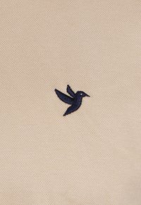 Beiger Stoff mit einem strukturierten Strickmuster, das ein kleines, navyblau gesticktes Vogel-Logo auf der linken Seite zeigt.