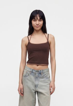 Ung kvinde med langt sort hår iført en brun cropped tanktop og løse lyseblå jeans, stående mod en ensfarvet hvid baggrund.