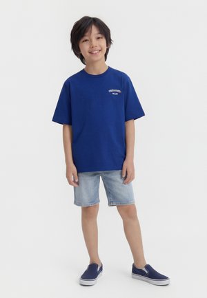 Usmievajúci sa chlapec stojaci v modrom tričku DSQUARED2, svetlých rifľových šortkách a tmavomodrých slip-on topánkach na bielom pozadí.