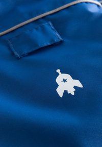 Blått tyg med en slät textur har en vit grafisk design som liknar en rymdraket med en stjärna. En liten rektangulär etikett är synlig.