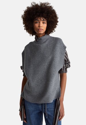 Frau mit lockigem Haar, die eine graue, ärmellose Strickweste über einem gestreiften Langarmshirt und blauen Jeans trägt, stehend vor weißem Hintergrund.