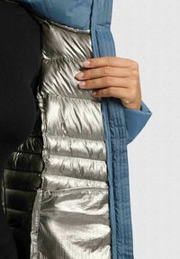 Giacca a vento blu con rivestimento isolante argentato metallico. L'interno presenta sezioni trapuntate e una tasca laterale. Una mano tiene l'apertura della giacca.