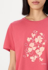 Kortärmad t-shirt med rund halsringning, tillverkad av mjukt rosa tyg som har en blomsterdesign i ljusrosa och vit kontur, logotyp i nedre hörnet.