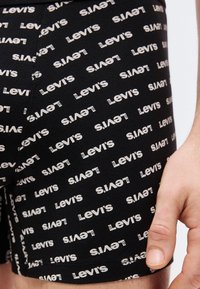 Κοντινό πλάνο μαύρων σορτς με λευκό επαναλαμβανόμενο μοτίβο λογότυπου Levi's και ορατό χέρι που ακουμπάει δίπλα στα σορτς.