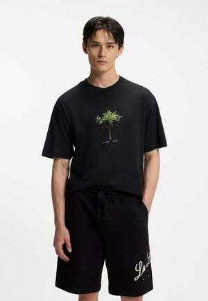Giovane uomo che indossa una t-shirt nera con grafica di palma e pantaloncini neri con scritta bianca, in piedi contro uno sfondo bianco uniforme.