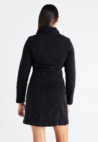 Μαύρο τζιν φόρεμα με μακριά μανίκια, εφαρμοστός μέσος και μήκος μέχρι το μέσο των μηρών. Διαθέτει γιακά και λεπτομέρεια με κορδόνι στην πλάτη για σμιλεμένο σχήμα.