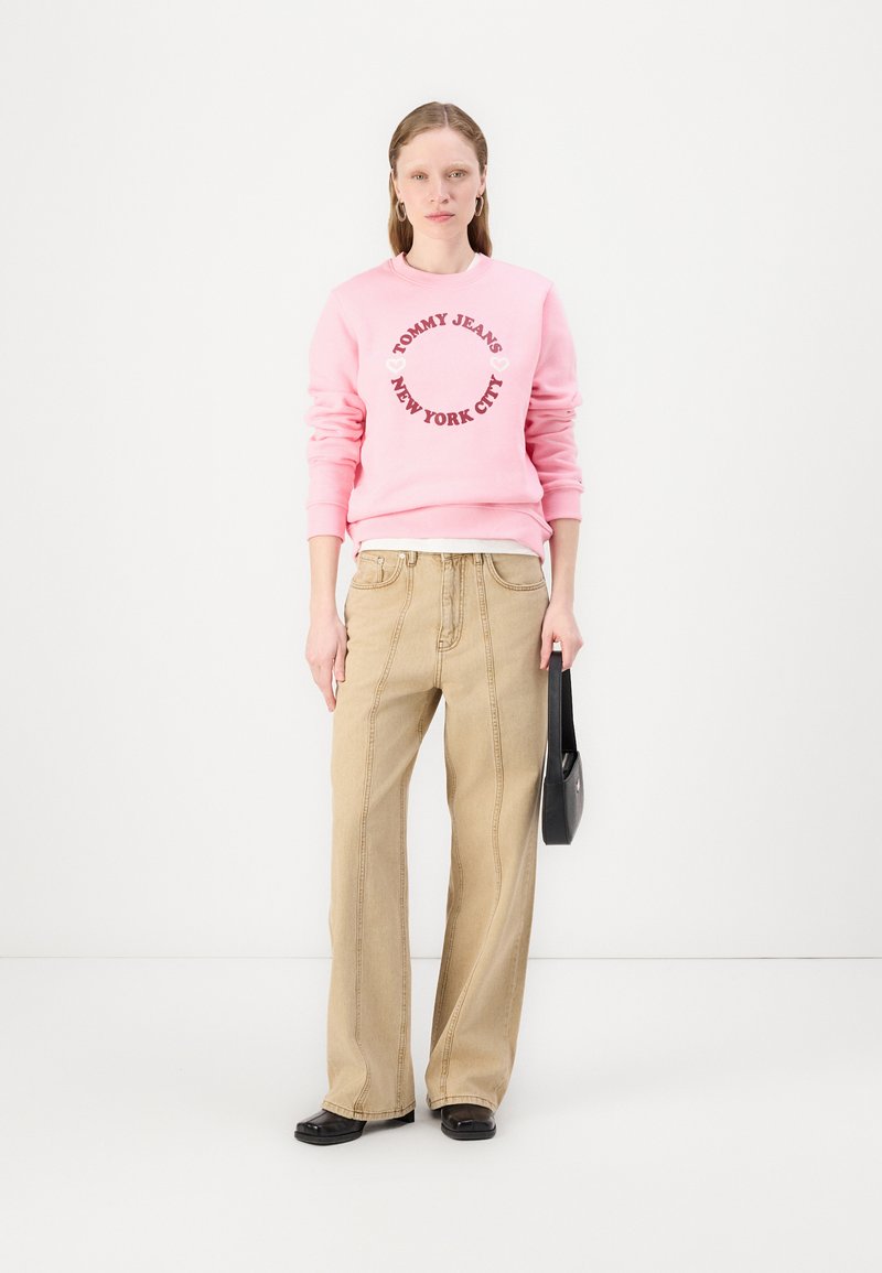 Felpa rosa con la stampa "TOMMY JEANS NEW YORK CITY" in rosso scuro, abbinata a pantaloni a gamba larga beige chiaro e scarpe nere basse.