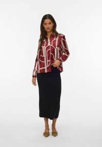 Camisa de satén rojo con patrones geométricos blancos, con botones en la parte delantera y mangas largas, combinada con una falda midi negra ajustada y zapatos puntiagudos de color verde oliva.