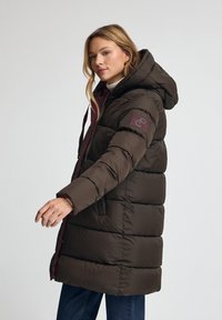 Brun pufferjacka med huva, med en quiltad design, ett vinrött foder och en logotyp patch på ärmen. Varm och isolerad.