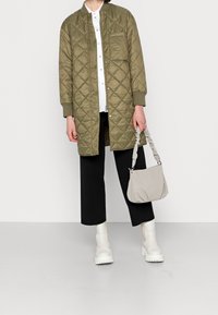 Abrigo largo acolchado verde con botones a presión, bolsillo frontal, combinado con una camisa blanca, pantalones anchos negros y botines en color blanco roto. Incluye un bolso gris.