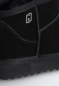 Bota de invierno de ante negro con suela gruesa, costuras visibles, forro interior suave y un pequeño logotipo metálico cerca del área del tobillo.