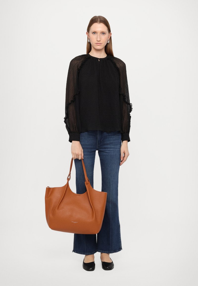 Sac fourre-tout en cuir marron avec une texture lisse et une ouverture sur le dessus. Modèle portant un blouse noire texturée, un jean évasé et des chaussures noires.