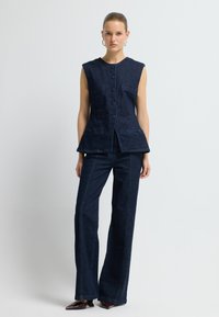 Tmavě modrá džínová bezrukávová košile s kulatým výstřihem, knoflíkovým zapínáním na přední straně a dvěma náprsními kapsami, doplněná o odpovídající rozšířené džíny.