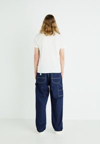 T-shirt bianco a maniche corte abbinato a pantaloni in denim blu scuro con vestibilità ampia, dotati di due grandi tasche laterali e cuciture a contrasto.