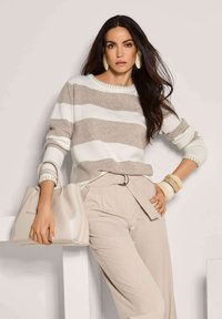 Frau in beigem und weiß gestreiften Pullover und beigen Hosen, hält eine beige Handtasche, trägt große Ohrringe und Armbänder.