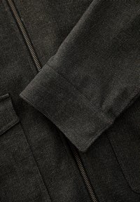 Teren textil gri închis, cu o textură vizibilă, fermoar și manșetă pliată. Materialul pare robust, având un model subtil și un finisaj neted.