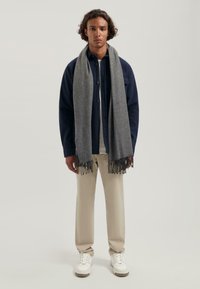 Grå tørklæde med frynser hængt om en models hals, parret med en marineblå skjorte med knapper og beige bukser, der har hvide sneakers.