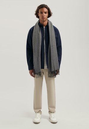 Grijze sjaal met franjes om de nek van een model, gecombineerd met een marineblauwe overhemd en beige broek, en witte sneakers.