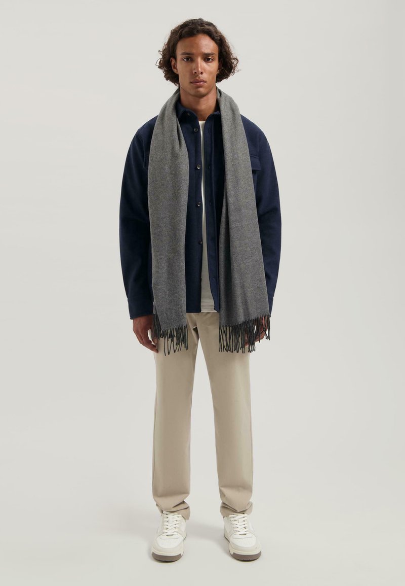 Grå tørklæde med frynser hængt om en models hals, parret med en marineblå skjorte med knapper og beige bukser, der har hvide sneakers.