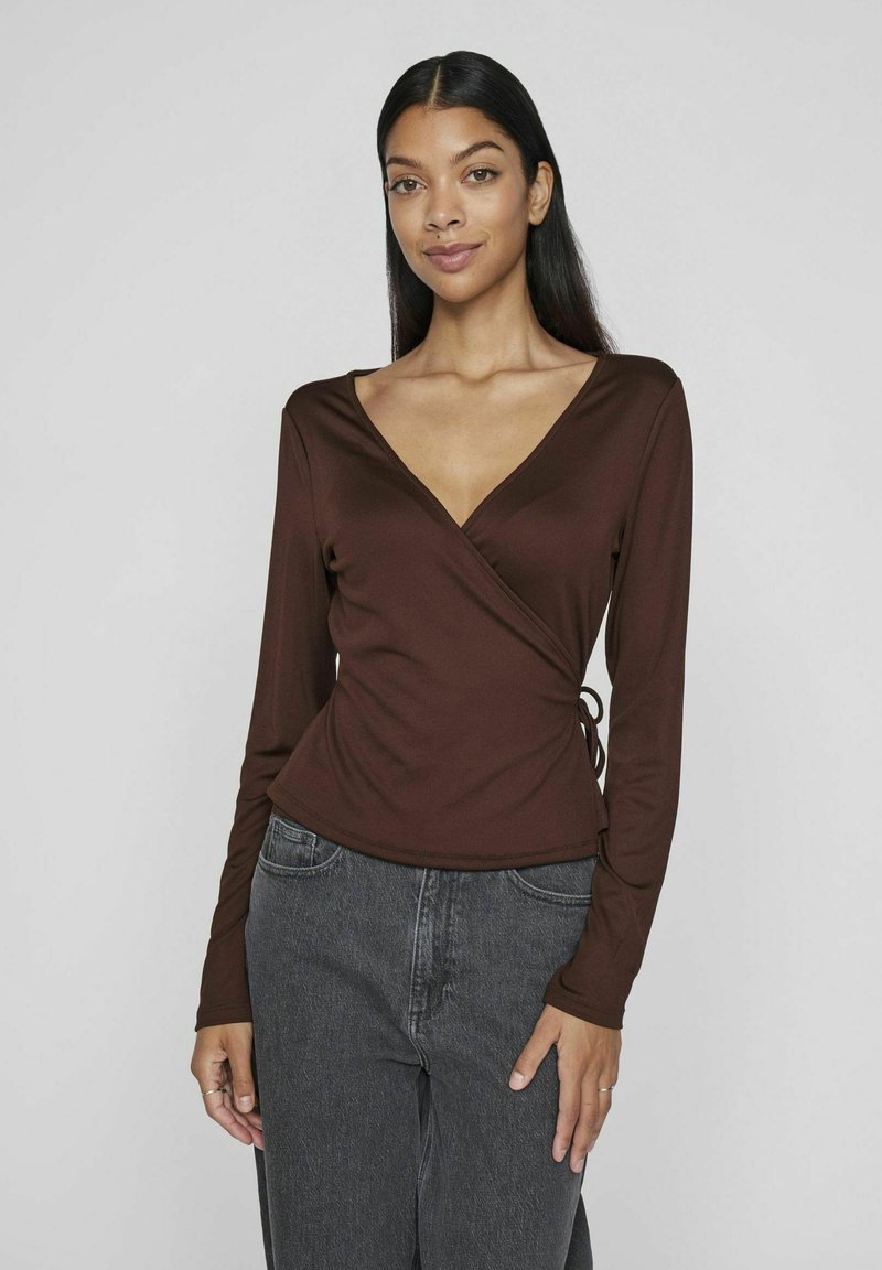 Top avvolgente marrone a maniche lunghe con scolla profonda a V, texture liscia, dettaglio annodato di lato, abbinato a jeans grigi a vita alta.
