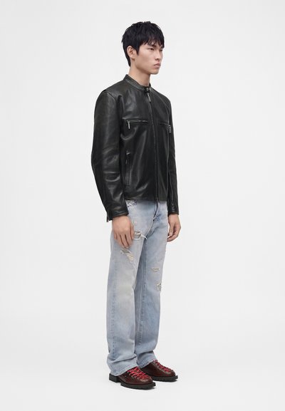 Homme portant une veste en cuir noire, un jean clair déchiré et des chaussures marron foncé avec des lacets rouges, debout devant un fond blanc uni.