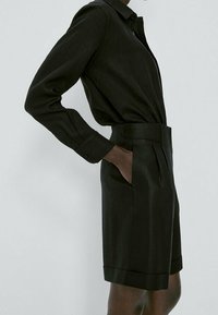 Personne portant une chemise noire à manches longues avec col, rentrée dans un short noir taille haute avec ceinture, les mains dans les poches, sur un fond uni.