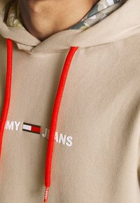 Sudadera con capucha beige con cordones rojos, con texto blanco "MY JEANS" y un pequeño logo con rayas horizontales. Con forro de tela con estampado floral.