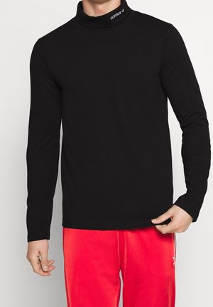Homme portant un col roulé noir à manches longues avec le logo "adidas" et un pantalon rouge vif, debout devant un fond blanc uni.